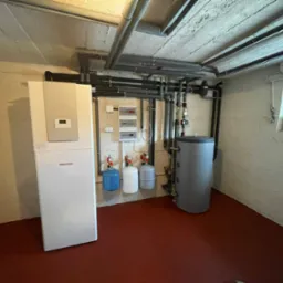 Kompaktowa kotłownia w piwnicy z piecem Stiebel Eltron, zbiornikiem akumulacyjnym, trzema małymi butlami ciśnieniowymi i rozbudowaną instalacją rur pod sufitem.