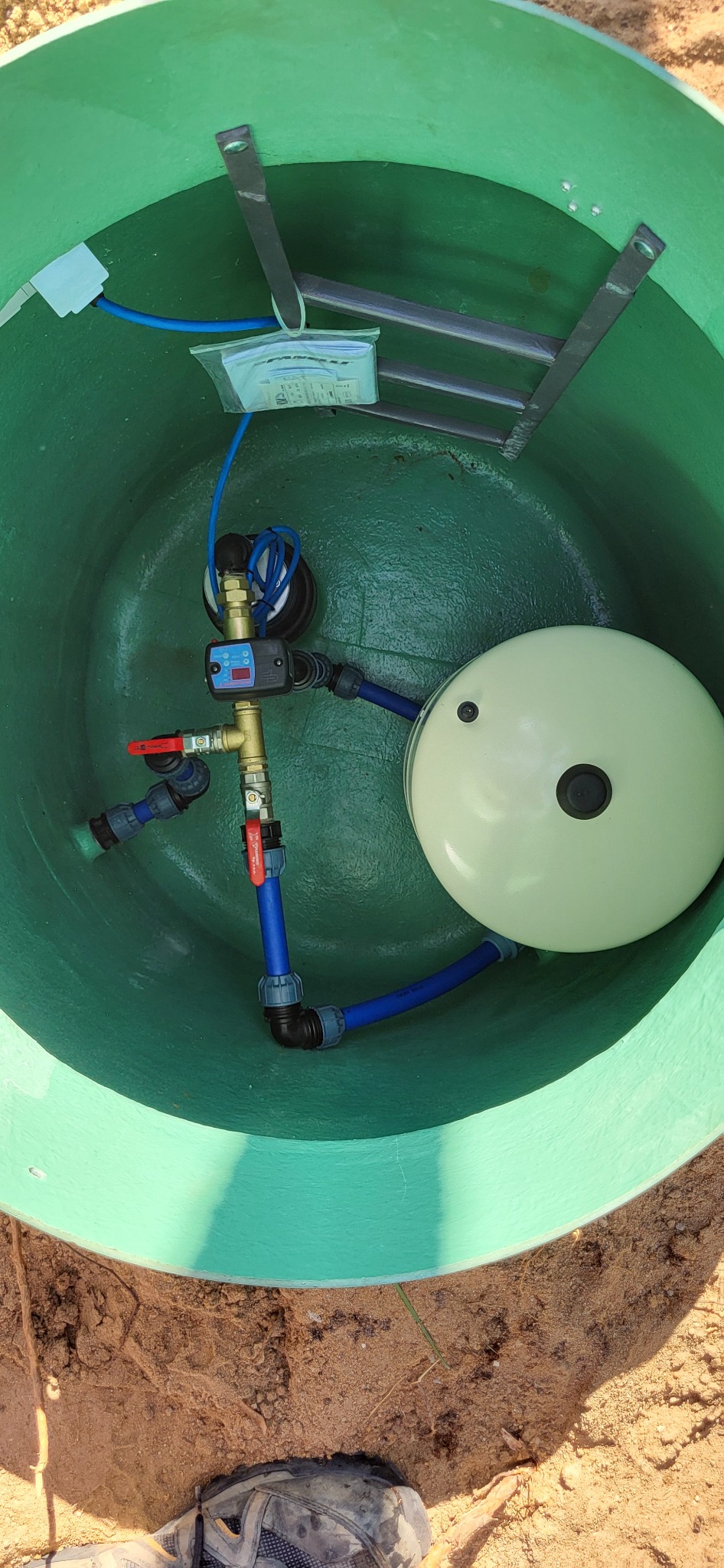 Wnętrze zielonej studni głębinowej z zainstalowanym osprzętem: zbiornik hydroforowy, manometr, zawory i rury z tworzywa sztucznego, drabinka w tle.