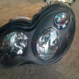 Czarny reflektor samochodowy, widoczne odbicia światła na kloszach, leżący na szorstkiej, betonowej powierzchni.