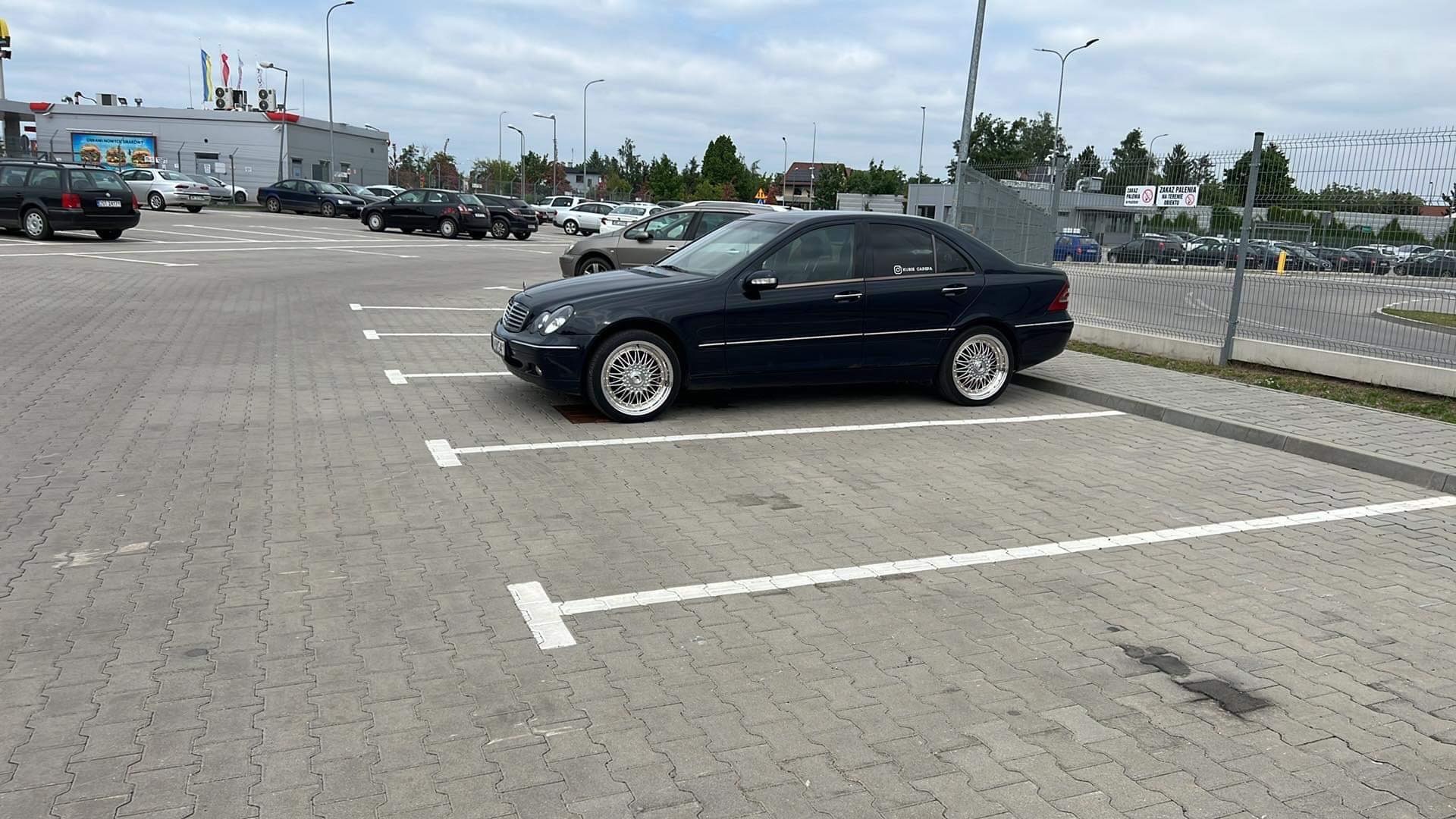 Granatowy samochód osobowy marki Mercedes zaparkowany na parkingu z kostki brukowej, z widocznymi chromowanymi felgami i naklejką na tylnej szybie.