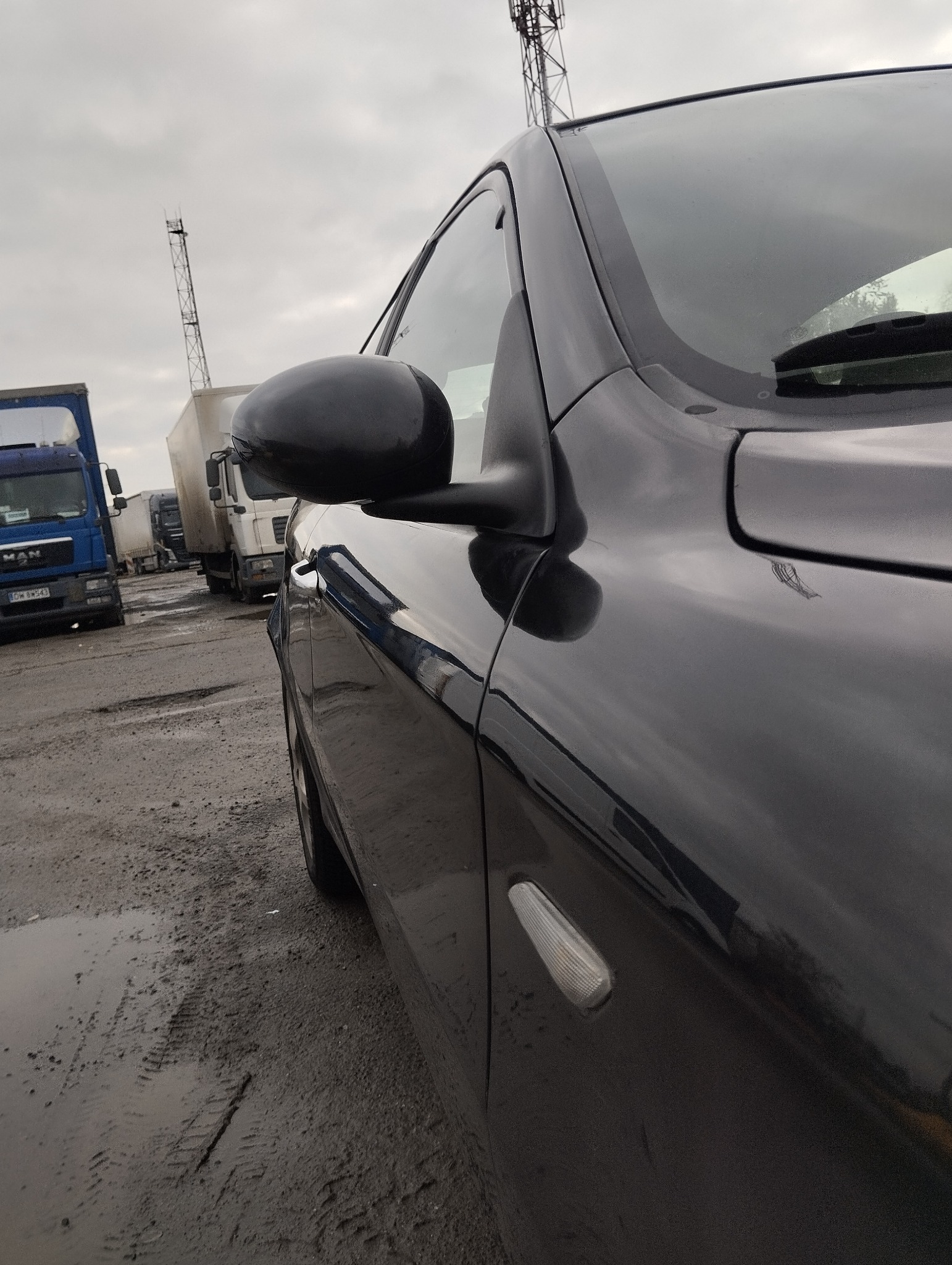 Błyszczący, czarny samochód na brudnym, przemysłowym parkingu z kałużami, widok z bliska na bok auta i lusterko, w tle zaparkowane ciężarówki i maszt telekomunikacyjny.