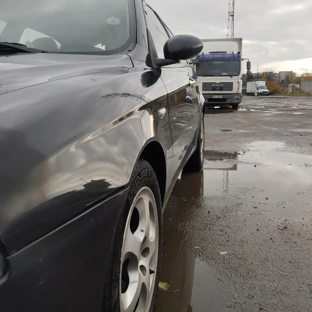 Ciemny samochód osobowy z widocznym uszkodzeniem lakieru na błotniku, stojący na mokrym placu z odbiciami, w tle zaparkowany biały samochód ciężarowy.