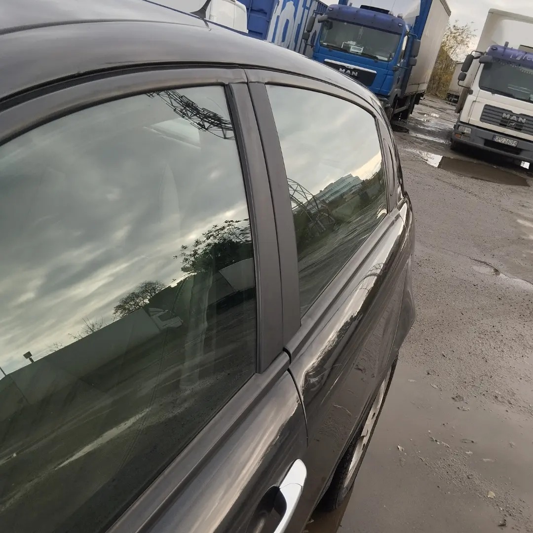 Ciemny samochód osobowy z widocznym odbiciem nieba i konstrukcji stalowej w szybie, stojący na mokrym placu z zaparkowanymi ciężarówkami w tle.