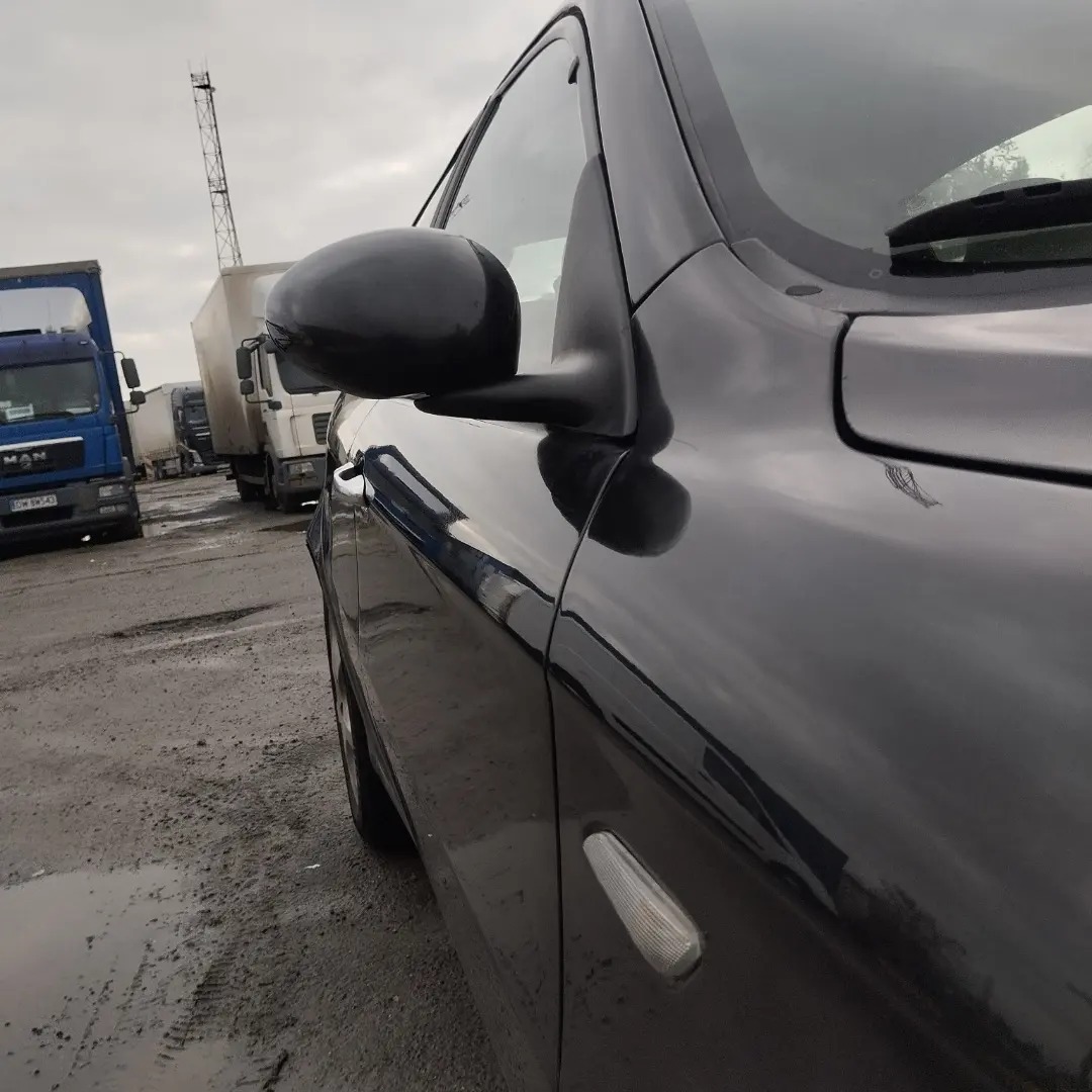 Błyszczący, czarny bok samochodu osobowego na tle placu z zaparkowanymi ciężarówkami i pochmurnym niebem.