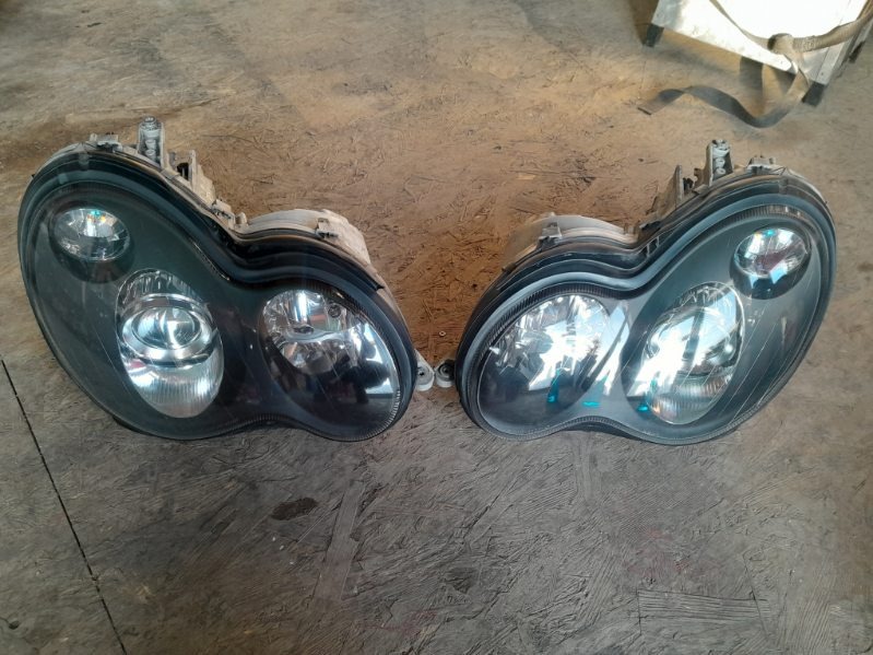 Dwie używane, czarne lampy samochodowe leżące na szarej, drewnianej powierzchni. Widoczne ślady użytkowania i brak czystości.