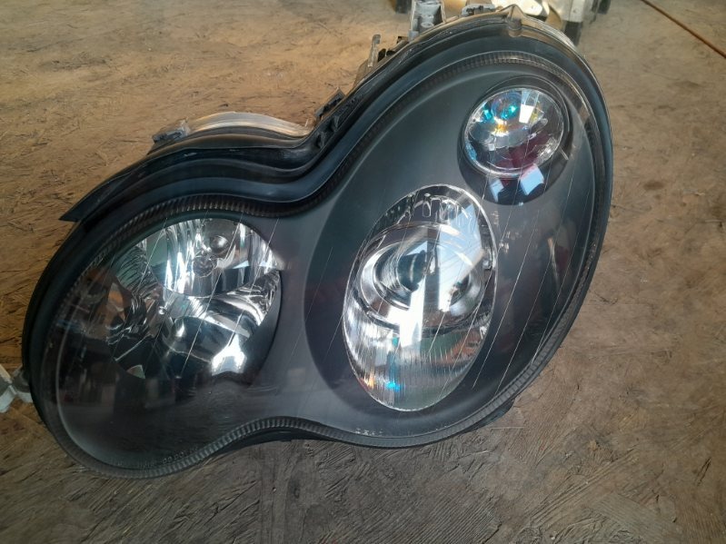 Czarny reflektor samochodowy, widoczne odbicia światła na kloszach, leżący na szorstkiej, betonowej powierzchni.