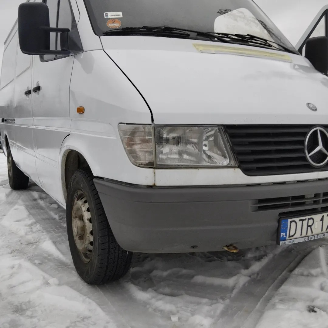 Biały samochód dostawczy Mercedes-Benz Sprinter na ośnieżonym terenie, widok z przodu na reflektor, koło i fragment zderzaka.
