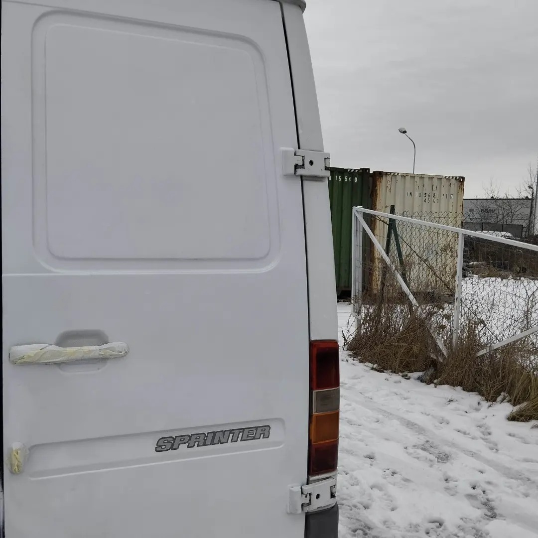 Tył białego samochodu dostawczego Sprinter z widocznymi uszkodzeniami taśmy na klamce, na tle zaśnieżonego terenu i kontenerów.