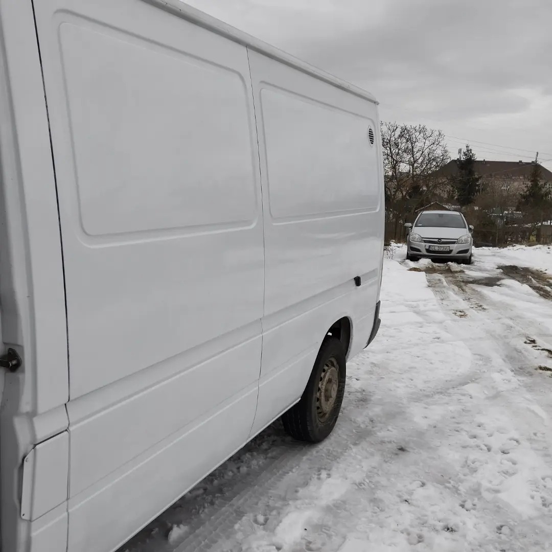 Biały furgon dostawczy z widocznym brudnym kołem na tle zaśnieżonego terenu i zaparkowanego samochodu osobowego.