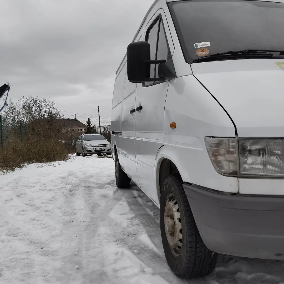 Biały samochód dostawczy zaparkowany na ośnieżonej drodze, widoczne ślady opon i inny samochód w tle, pochmurne niebo.