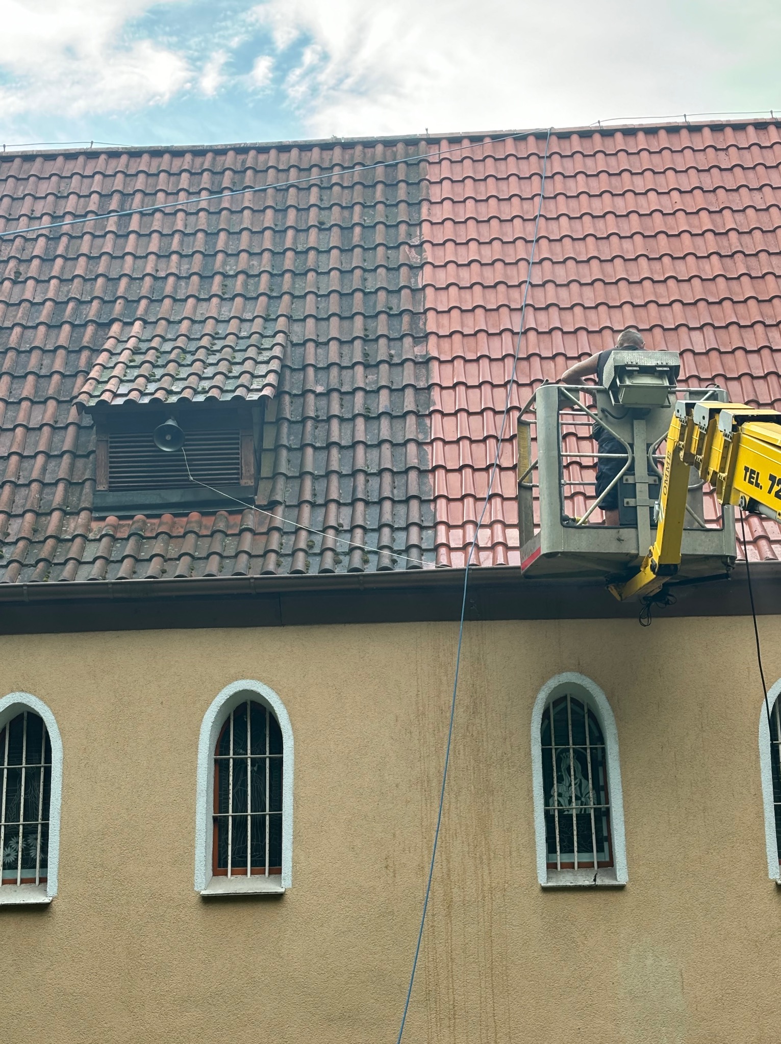 Czyszczenie połowy dachu z czerwonej dachówki z mchu i brudu za pomocą podnośnika koszowego na tle beżowej elewacji budynku z oknami z kratami.