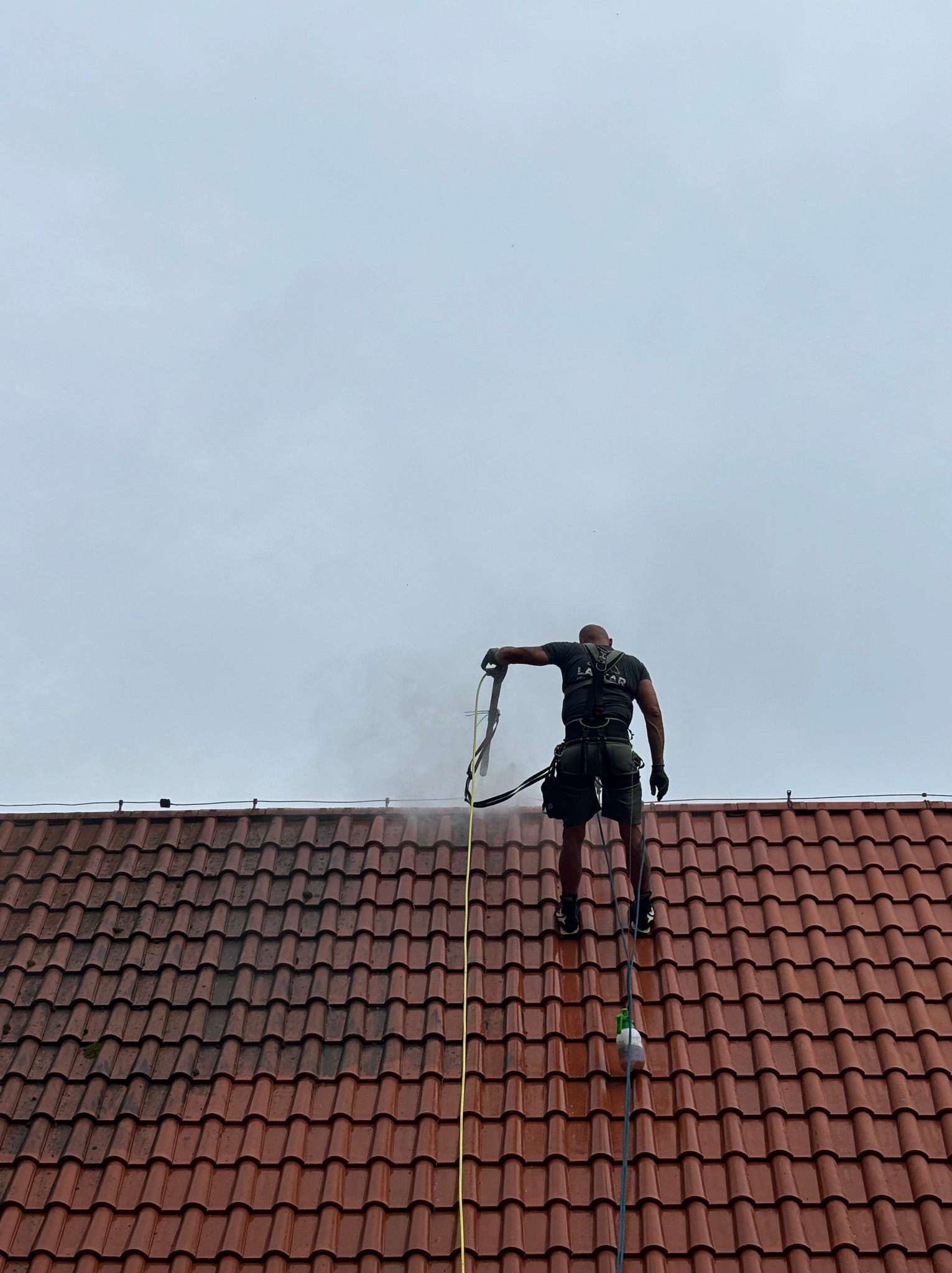 Pracownik w uprzęży, od tyłu, myje ciśnieniowo czerwoną dachówkę, widoczny efekt czyszczenia i para wodna.