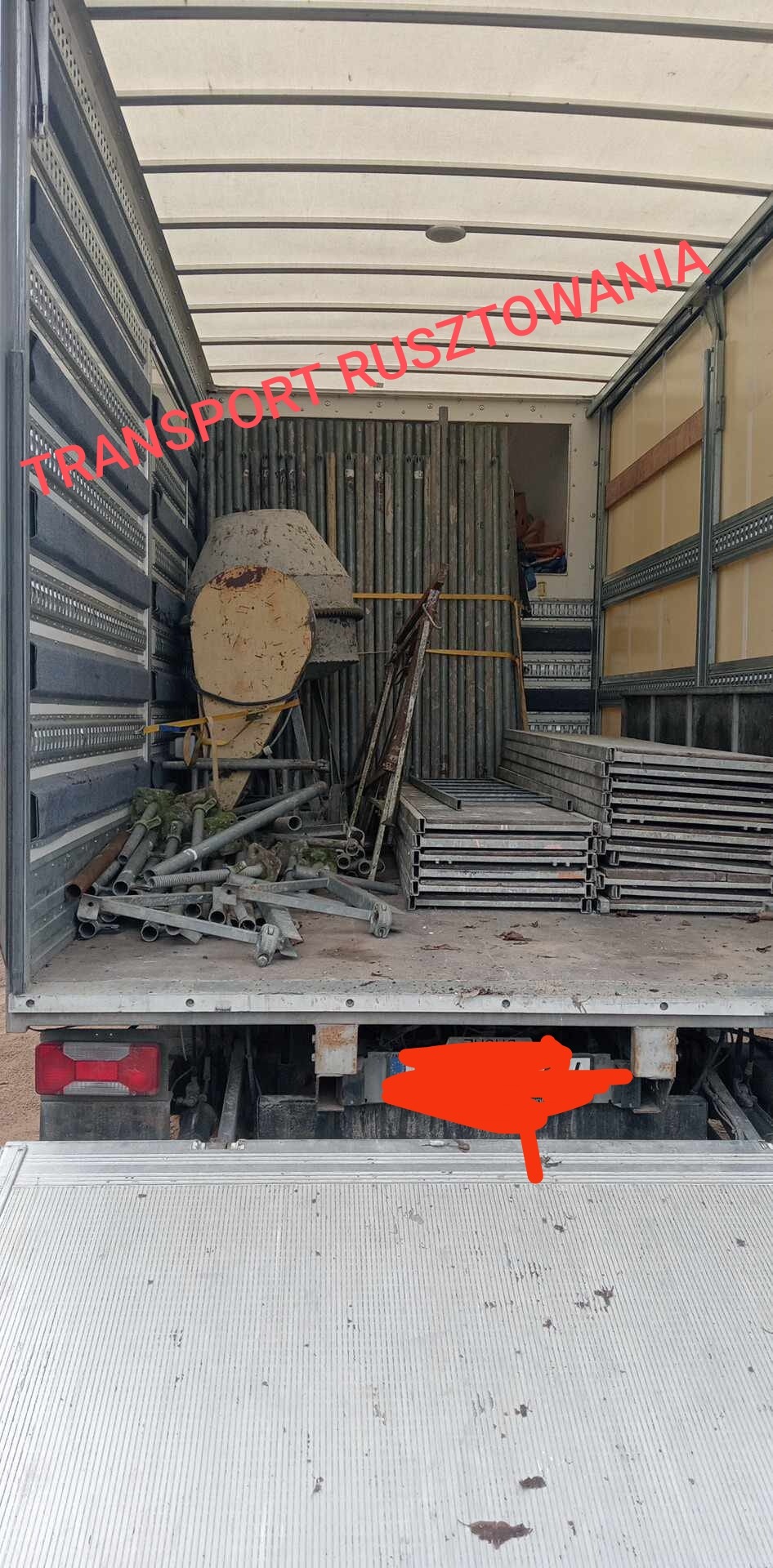 Wnętrze skrzyni ładunkowej samochodu ciężarowego wypełnione elementami rusztowania, w tym drabiny, rury i podesty, oraz starą betoniarką. Widoczne ślady użytkowania i zabrudzenia.