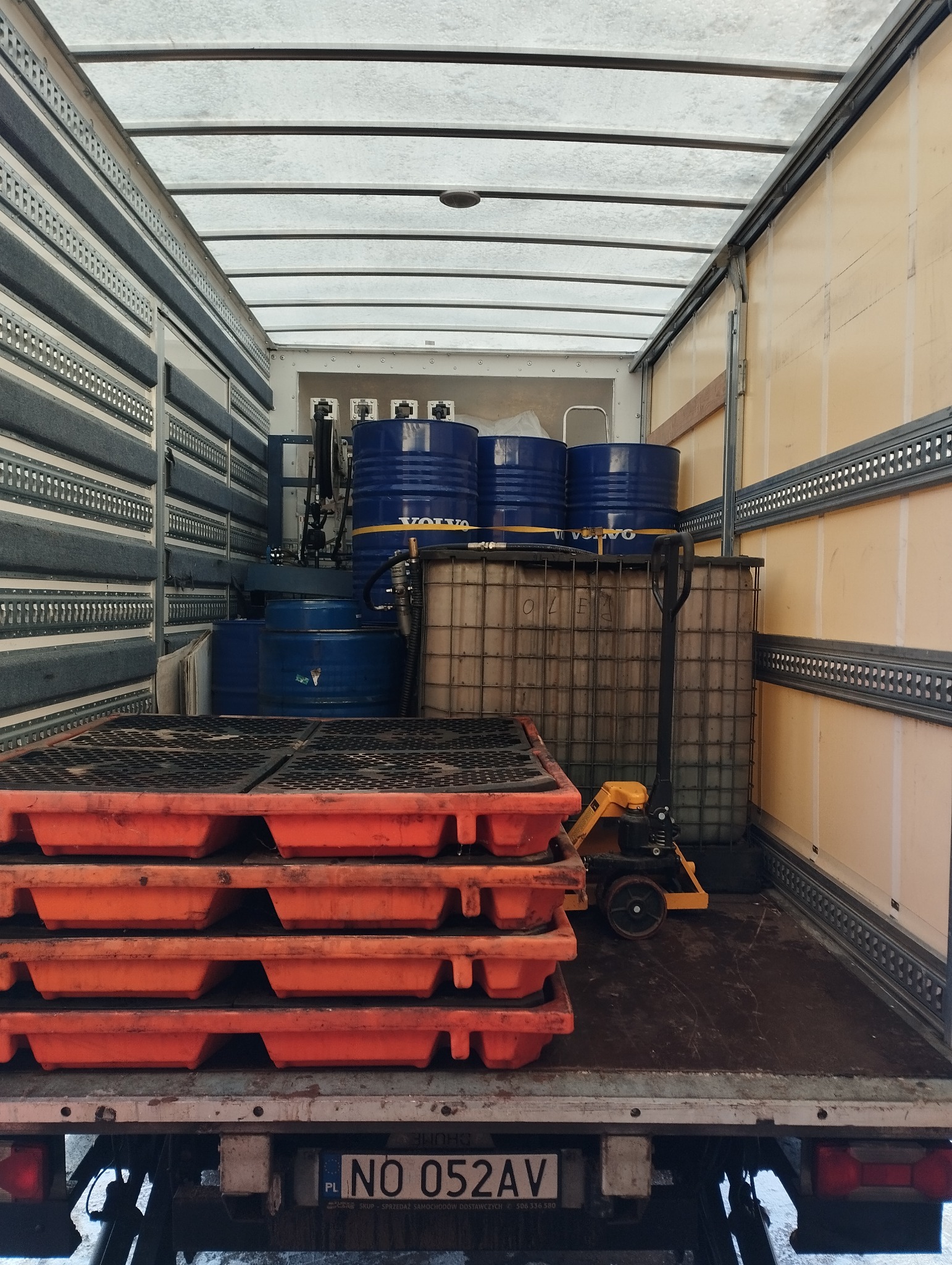Wnętrze przestrzeni ładunkowej samochodu ciężarowego wypełnione niebieskimi beczkami z logo Volvo, paletopojemnikiem IBC z napisem OLEJ, paletami ociekowymi i wózkiem paletowym.