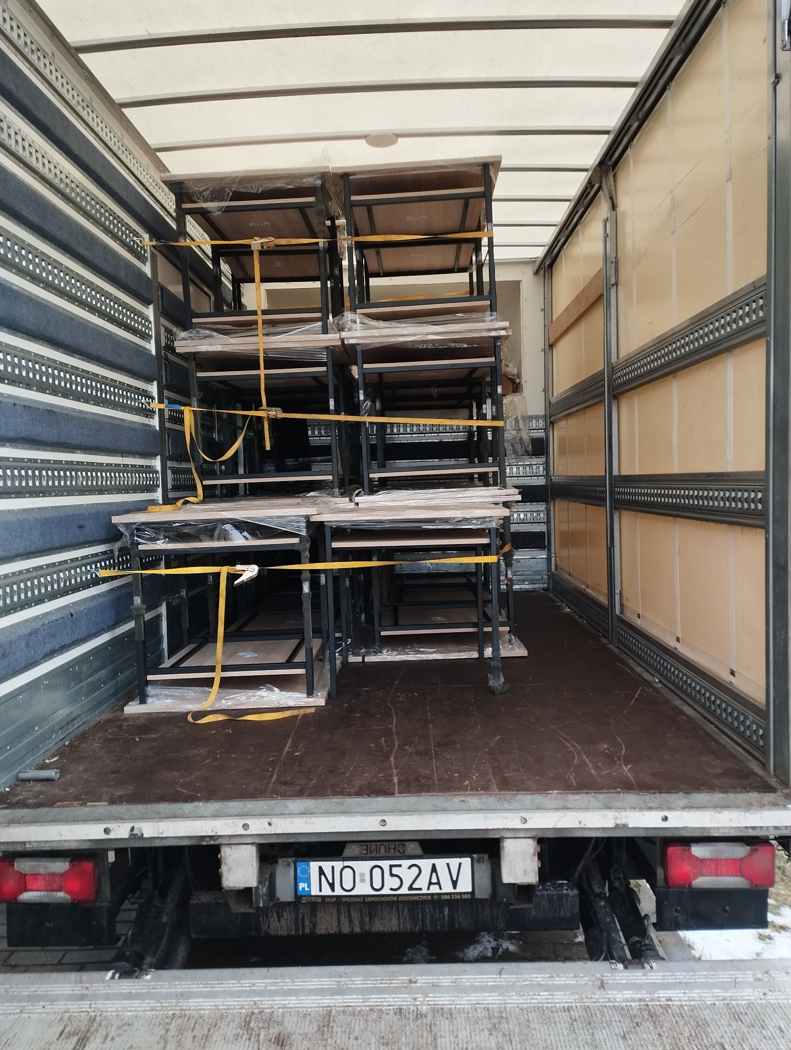 Wnętrze ciężarówki z załadowanymi i zabezpieczonymi pasami regałami z drewnianymi półkami, owiniętymi folią stretch.