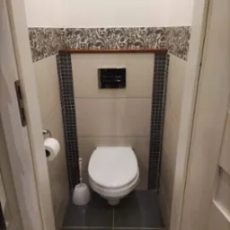 Kompaktowa toaleta w wykończonej łazience, z mozaiką w odcieniach szarości i beżu, nad którą umieszczono dekoracyjny pas z motywem roślinnym i drewnianą półką.