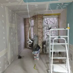 Pokój w trakcie prac wykończeniowych: ściany obłożone płytami kartonowo-gipsowymi, częściowo zaszpachlowane, widoczne kable zwisające z sufitu, drabina aluminiowa i sterta worków z zaprawą...