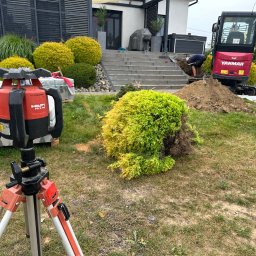 HYDROTECH - Czerwony niwelator laserowy Hilti na trójnogu na trawniku, w tle koparka Yanmar, pracownik przy wykopie, dom z szarymi schodami i ozdobne krzewy.