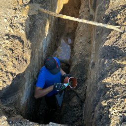 HYDROTECH - Pracownik w wykopie łączy rury kanalizacyjne, widoczna woda gruntowa i łopata oparta o ścianę wykopu.