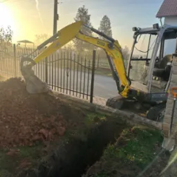 Żółta mini koparka Wacker Neuson ET18 przy wykopie wąskiego rowu w ziemi, obok metalowego ogrodzenia i chodnika. W tle dom i poranne słońce.