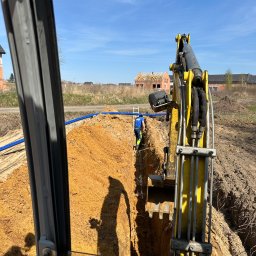 HYDROTECH - Widok z kabiny koparki na wykop z ułożonymi niebieskimi rurami, pracownik w oddali w odblaskowym ubraniu, w tle budowa domu w stanie surowym.
