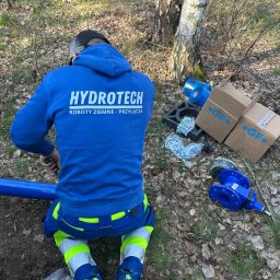 HYDROTECH - Monter w niebieskiej bluzie z napisem HYDROTECH na plecach, łączy niebieską rurę z użyciem narzędzi, w tle kartony z logo +GF+ i leżące elementy instalacji na ziemi pokrytej liśćmi.