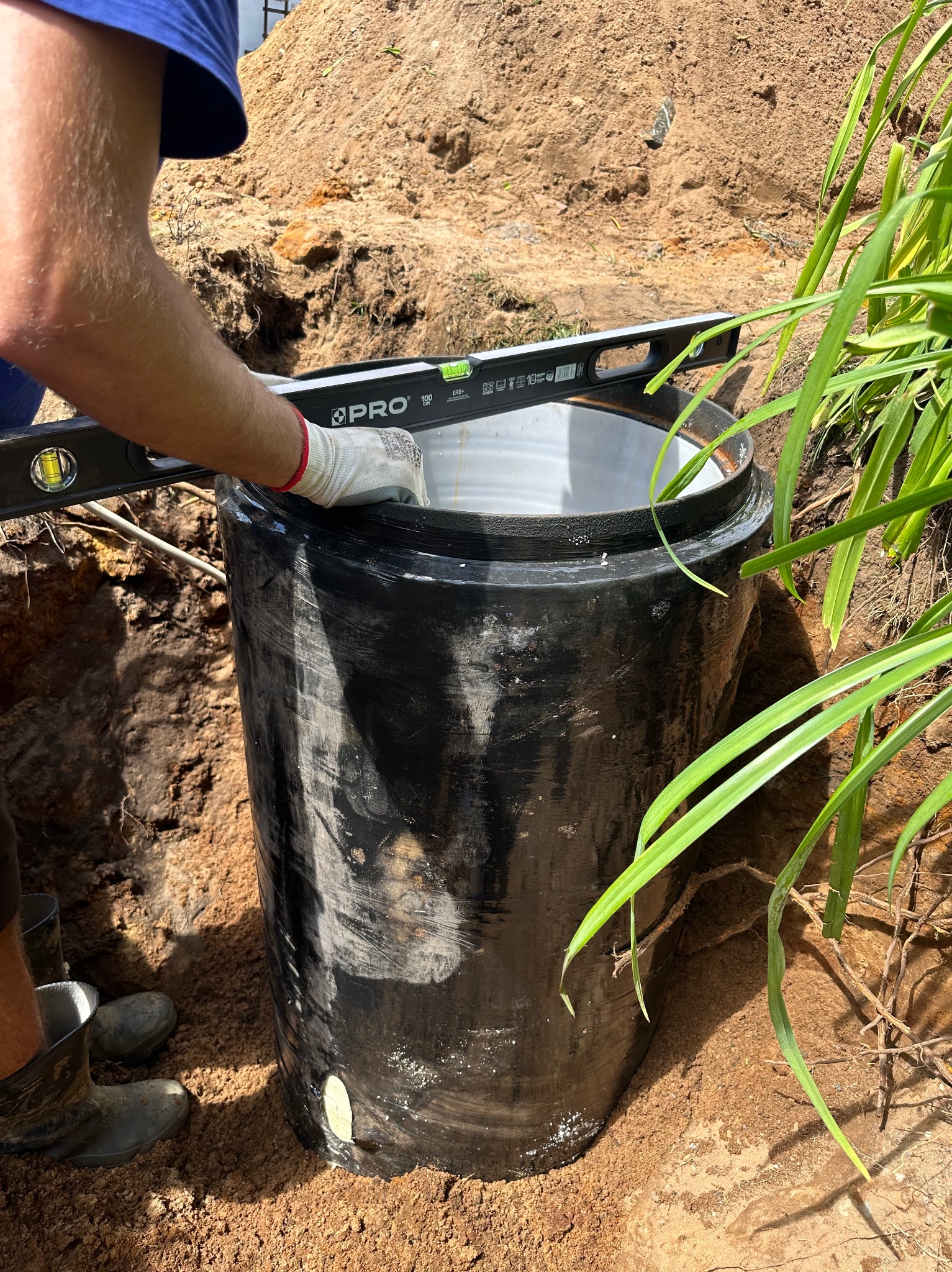 Czarna, cylindryczna studzienka z tworzywa sztucznego, częściowo zagłębiona w wykopie z piasku, poziomowana przez pracownika w rękawicach roboczych za pomocą poziomicy PRO. Obok rosną zielone rośliny.