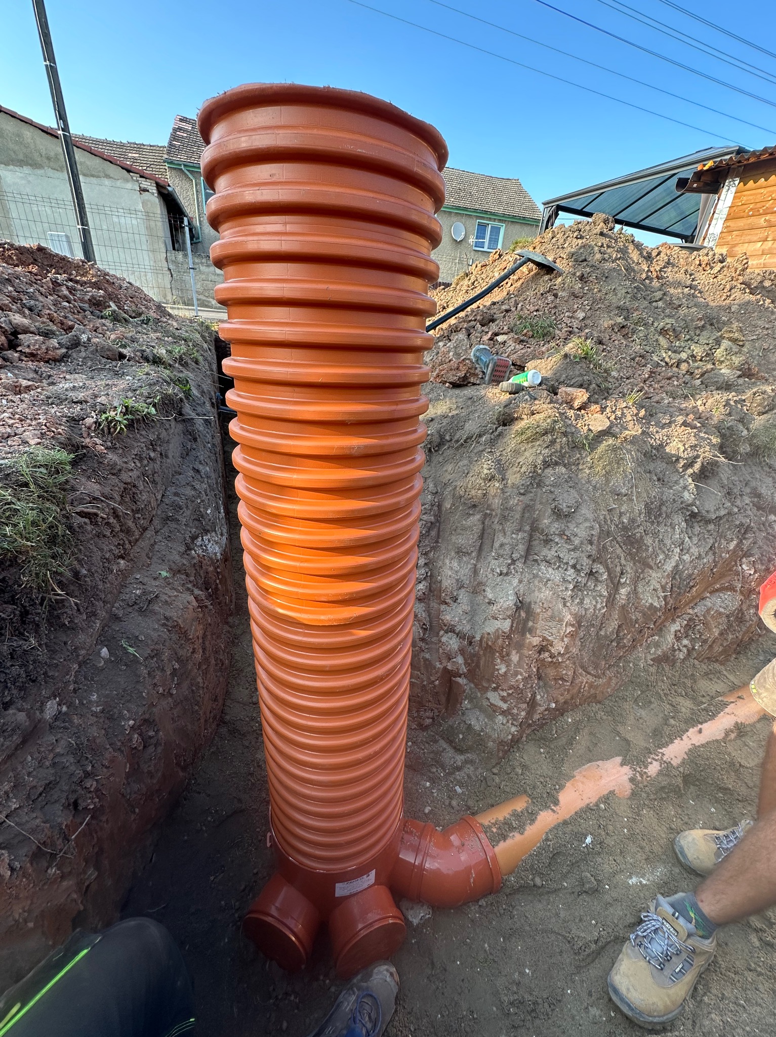 Pomarańczowa, karbowana rura pionowa z podłączeniami w wykopie ziemnym, widoczne fragmenty butów i spodni osoby wykonującej prace, w tle zabudowania.