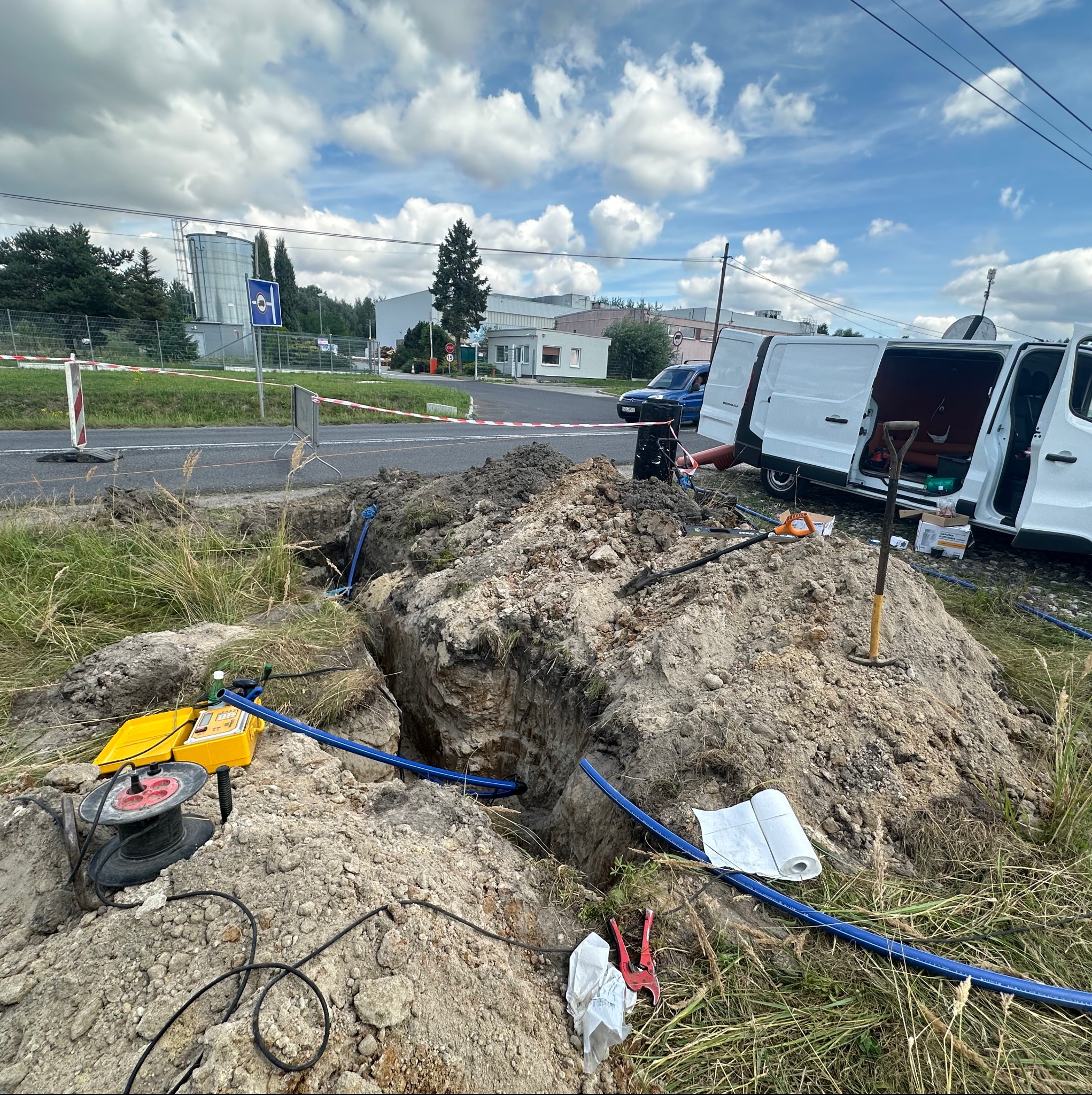 Głęboki wykop w ziemi z niebieskimi rurami, żółty miernik i zwinięty kabel na ziemi, obok leżą czerwone nożyce do rur i notatnik, w tle zaparkowane białe busy i budynki przemysłowe pod błękitnym...