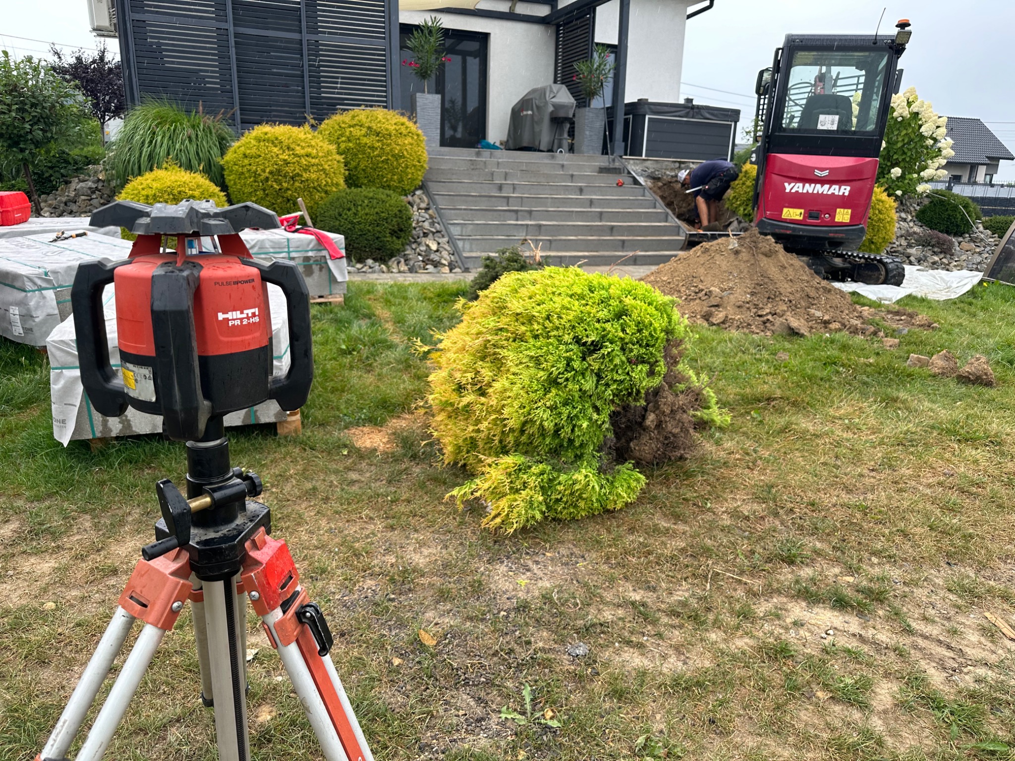 Czerwony niwelator laserowy Hilti na trójnogu na trawniku, w tle koparka Yanmar, pracownik przy wykopie, dom z szarymi schodami i ozdobne krzewy.