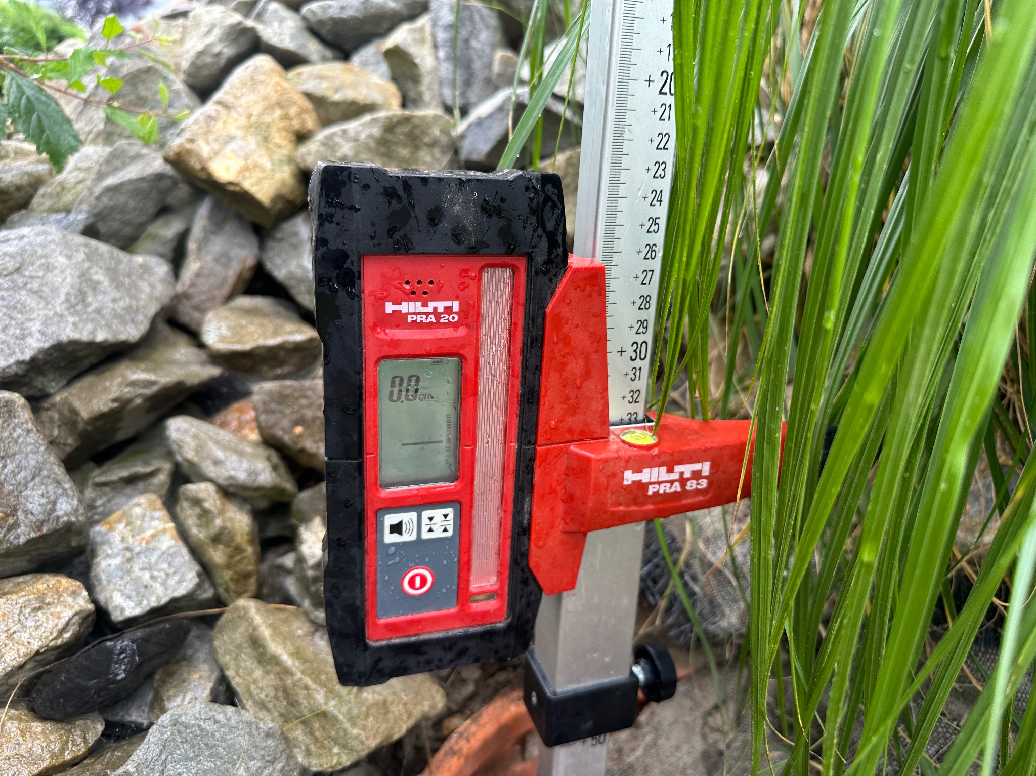 Hilti PRA 20 laserowy odbiornik i Hilti PRA 83 łata pomiarowa ustawione na tle kamieni i zielonej trawy, wskazujące poziom gruntu.