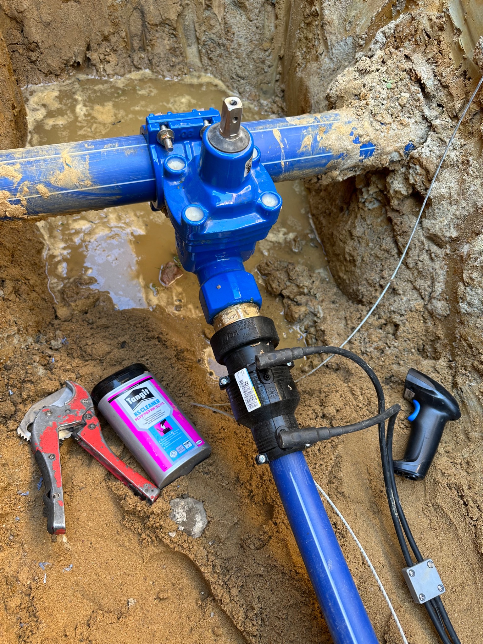 Niebieska rura wodociągowa z zaworem, umieszczona w wykopie wypełnionym wodą, obok leżą obcinak do rur, środek czyszczący Tangit KS Cleaner oraz skaner kodów kreskowych.