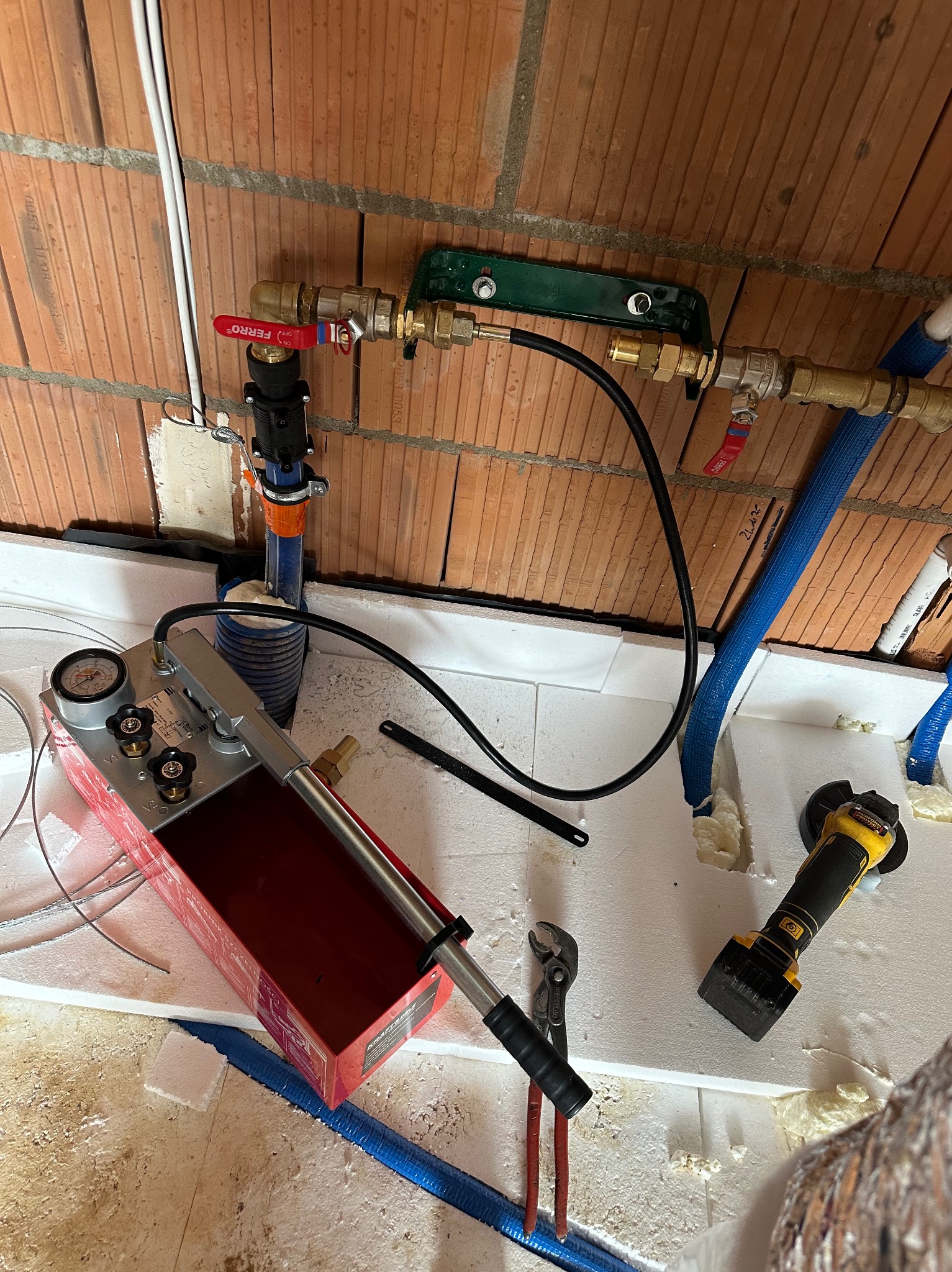 Ciśnieniowa próba instalacji wodnej z użyciem pompy ręcznej i manometru, widoczne rury miedziane z zaworami Fererro, niebieskie rury w izolacji, szlifierka kątowa i kombinerki na styropianie