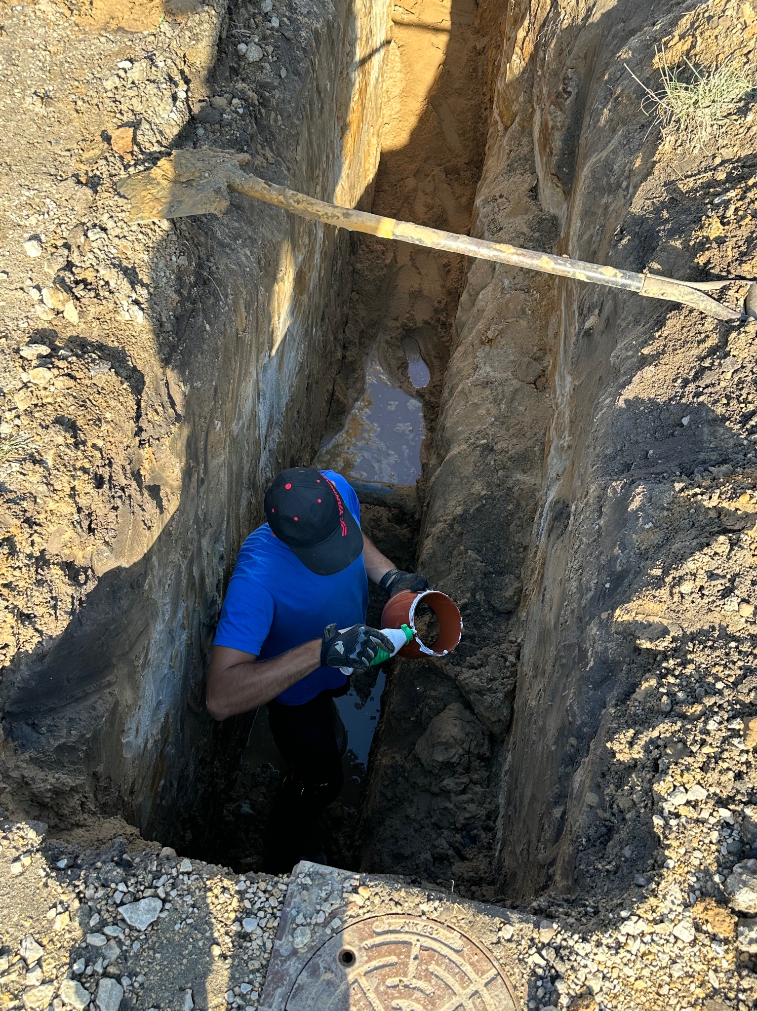 Pracownik w wykopie łączy rury kanalizacyjne, widoczna woda gruntowa i łopata oparta o ścianę wykopu.