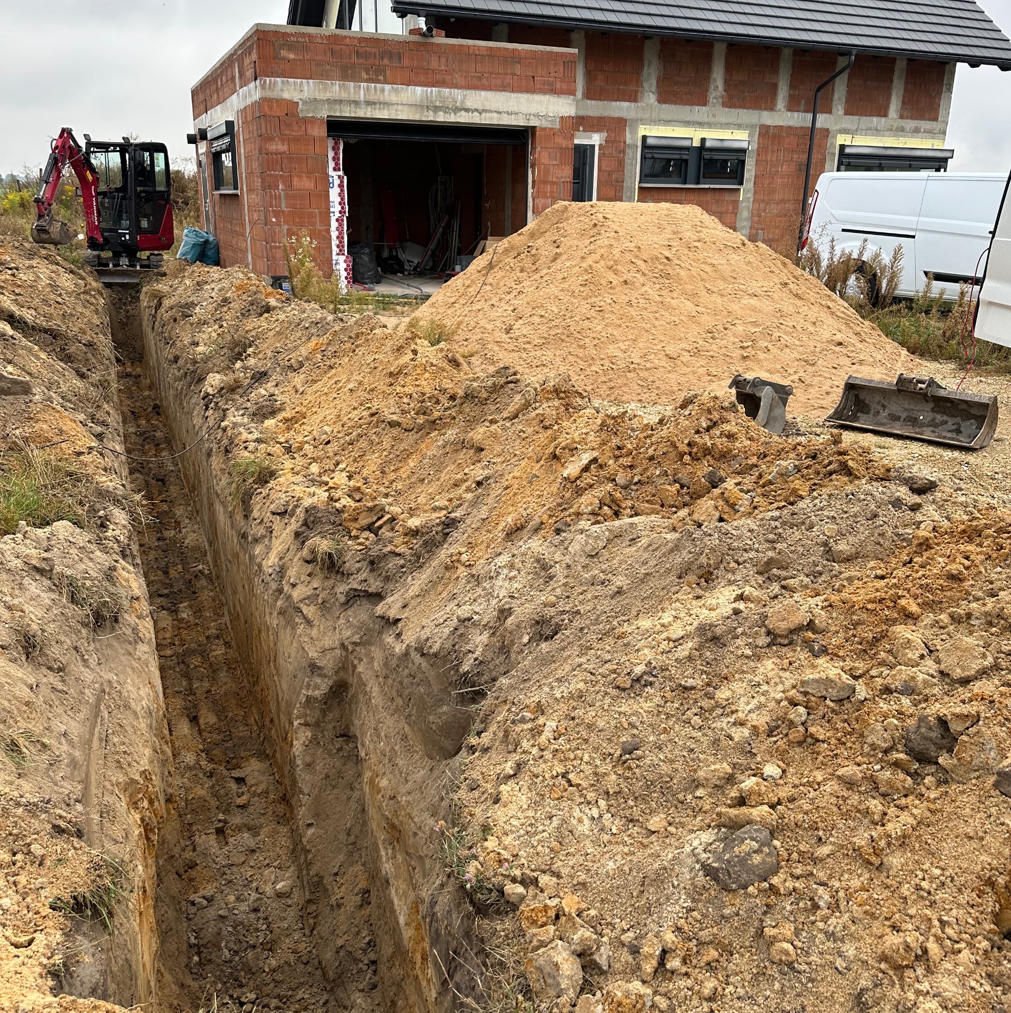 Wąski wykop w ziemi, z małą koparką na końcu, obok sterta piasku i budynek w trakcie budowy z białym vanem w tle.