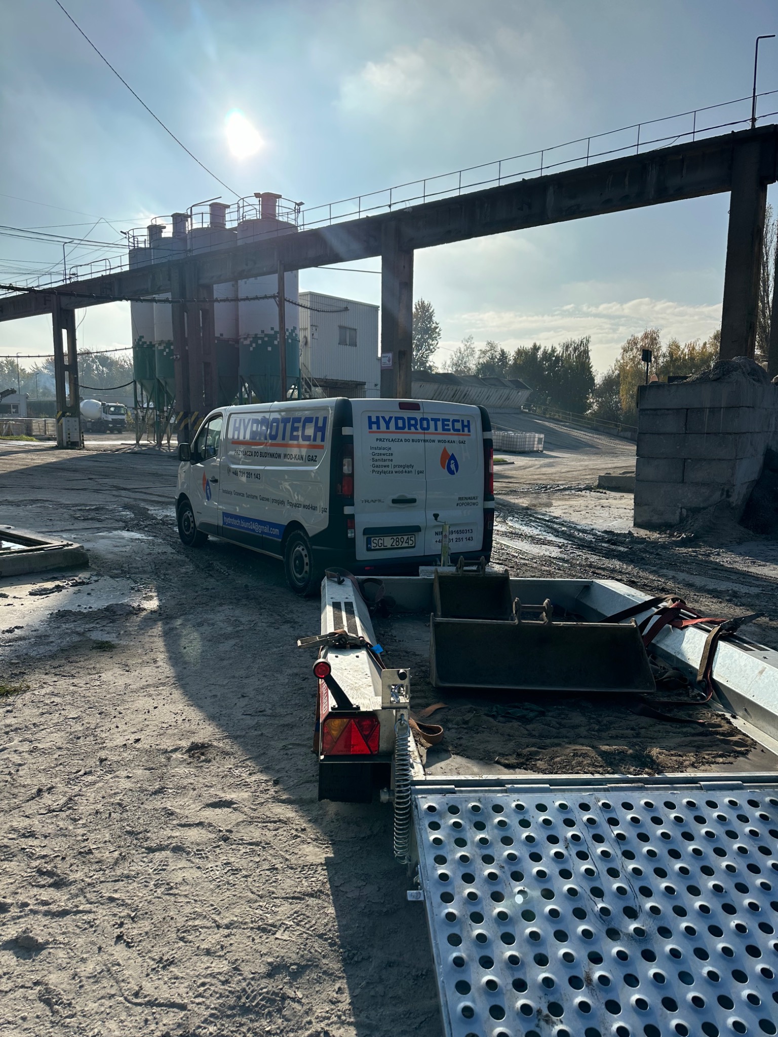 Furgonetka firmy Hydrotech z przyczepą i narzędziami na placu budowy, w tle silosy i konstrukcja mostowa pod słońce.