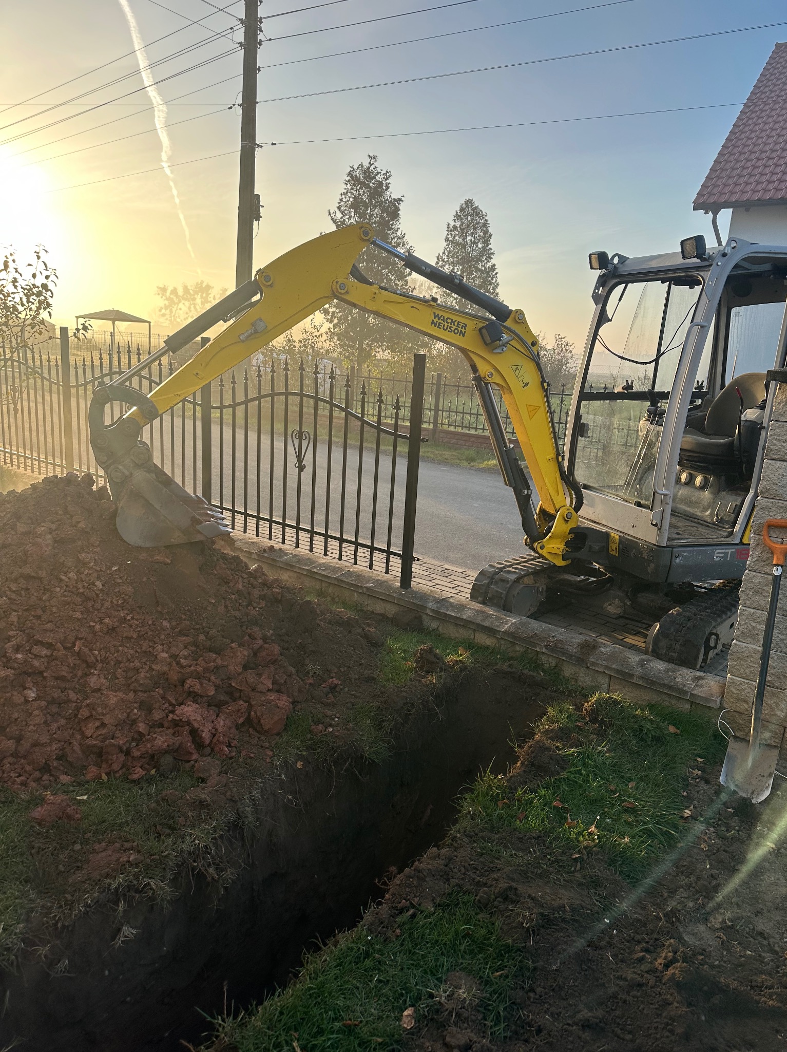 Żółta mini koparka Wacker Neuson ET18 przy wykopie wąskiego rowu w ziemi, obok metalowego ogrodzenia i chodnika. W tle dom i poranne słońce.