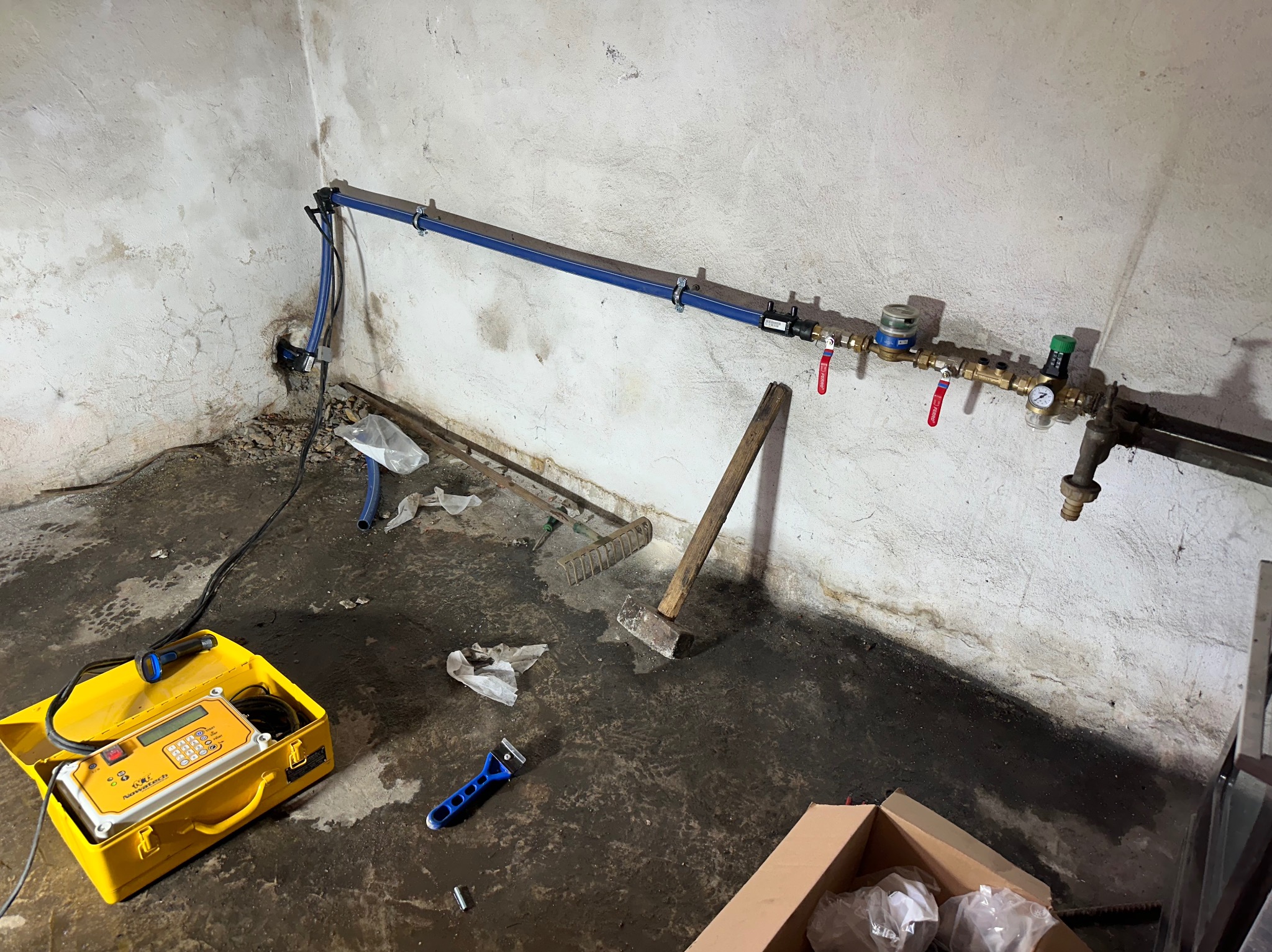 Nowe rury wodociągowe z licznikiem i zaworami zamontowane w piwnicy, widoczny sprzęt pomiarowy w żółtej walizce i narzędzia na brudnej podłodze.