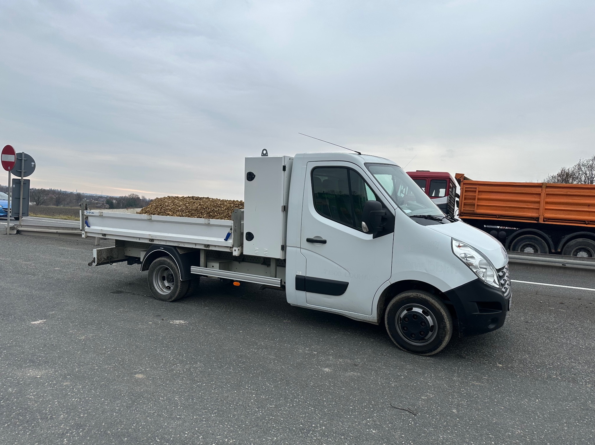 Biała ciężarówka z otwartą skrzynią załadowana ziemią lub gruzem, stojąca na asfalcie, z pomarańczową ciężarówką w tle i znakami drogowymi po lewej stronie.