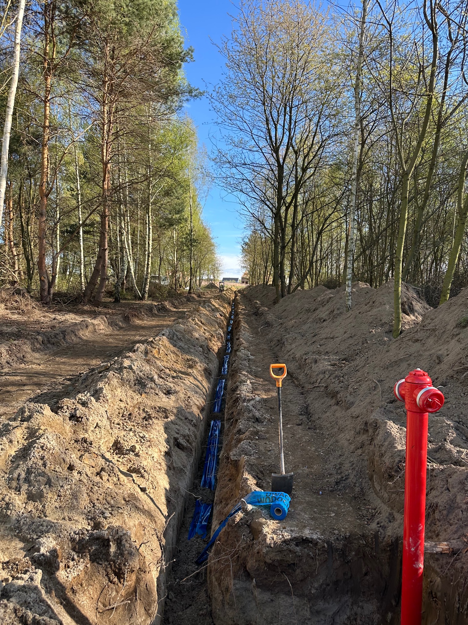 Wykop w ziemi z ułożonymi rurami w niebieskiej folii ochronnej, widoczna łopata z pomarańczowym uchwytem i czerwony hydrant, drzewa w tle.