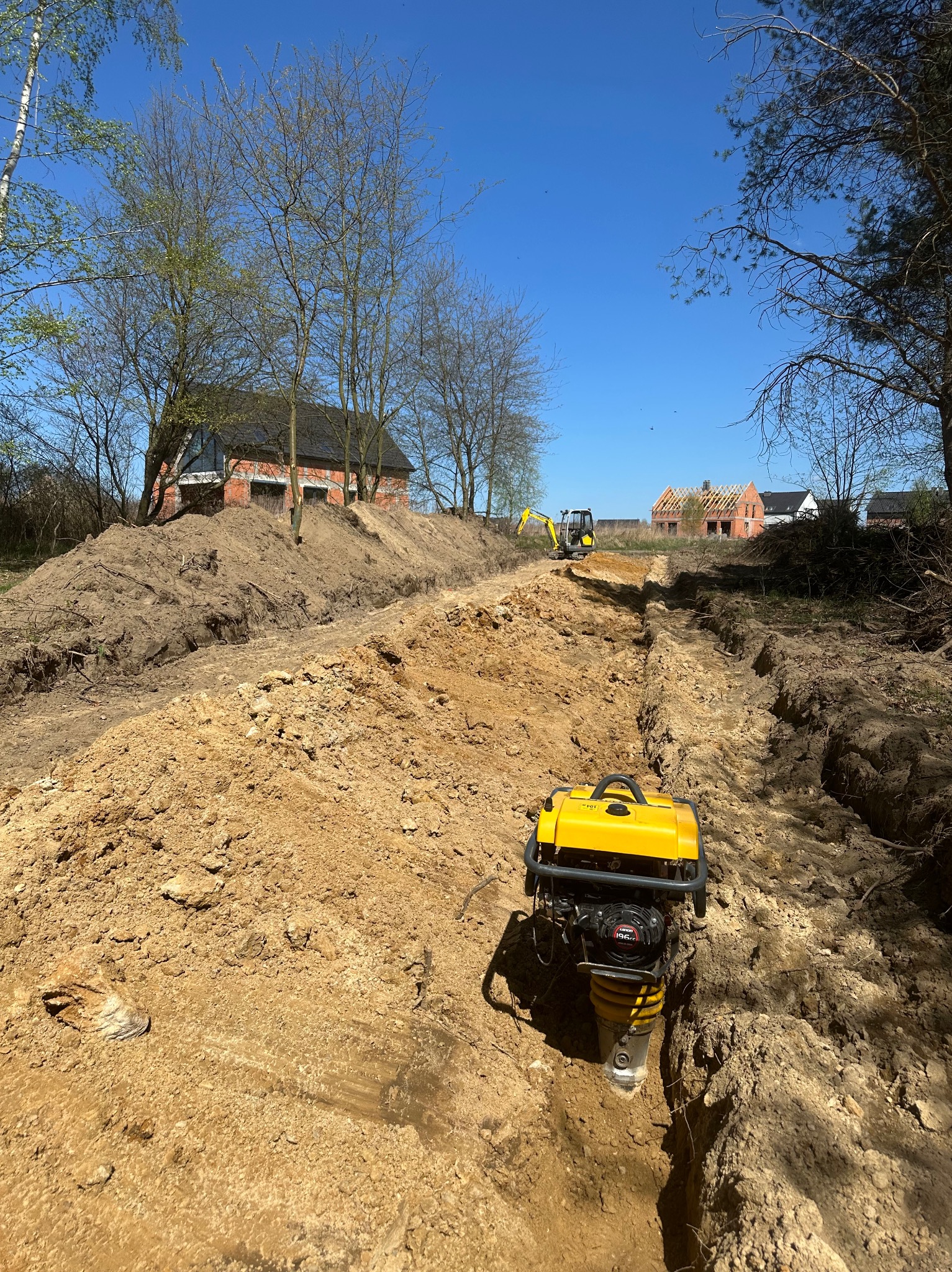 Żółta ubijarka stopowa zagęszcza ziemię w wykopie pod instalacje, w tle minikoparka i budynki w stanie surowym otwarte, błękitne niebo.