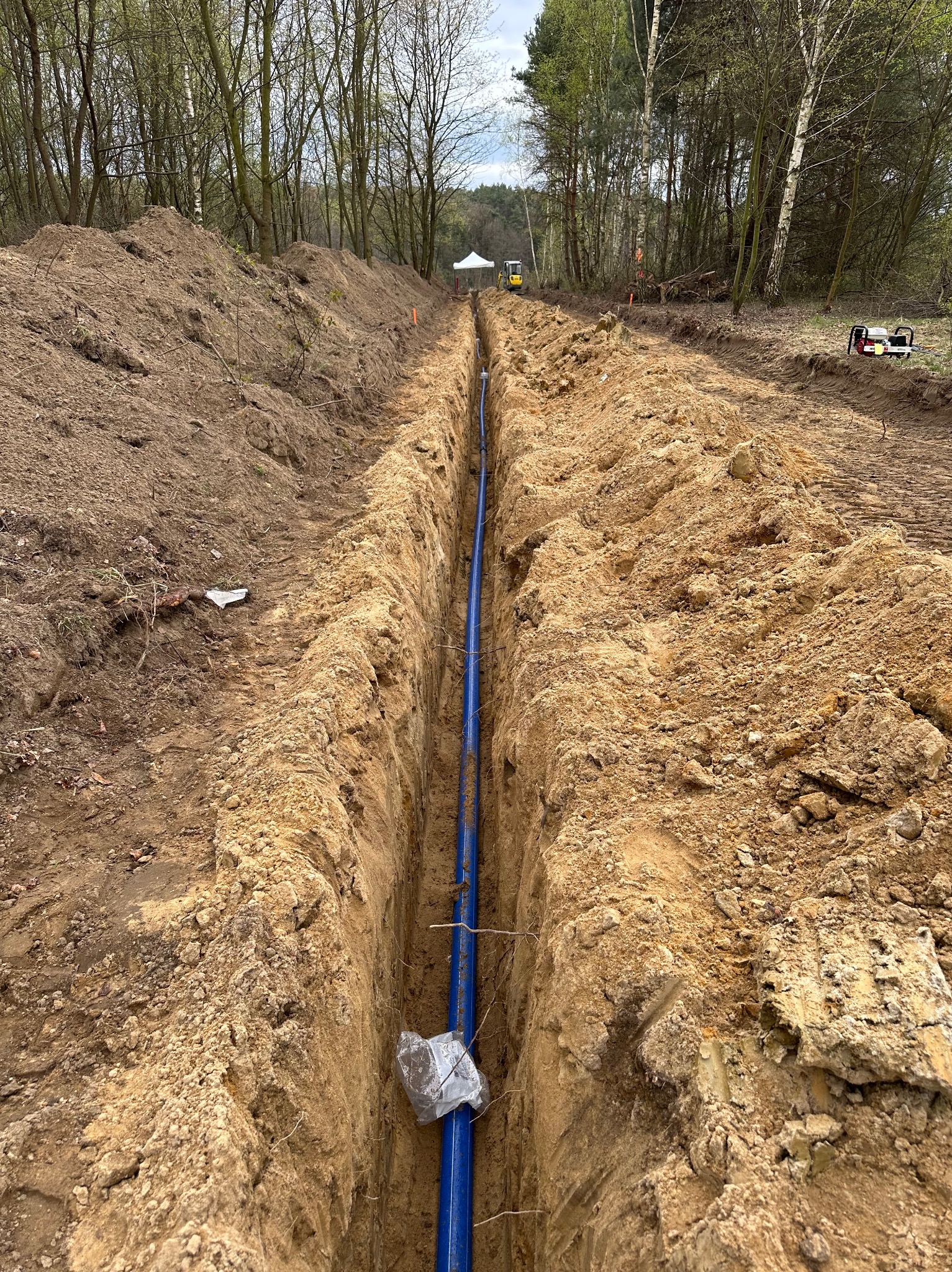 Wykopany wąski rów w ziemi z ułożoną niebieską rurą, widoczny sprzęt budowlany w oddali i usypana ziemia po bokach rowu.