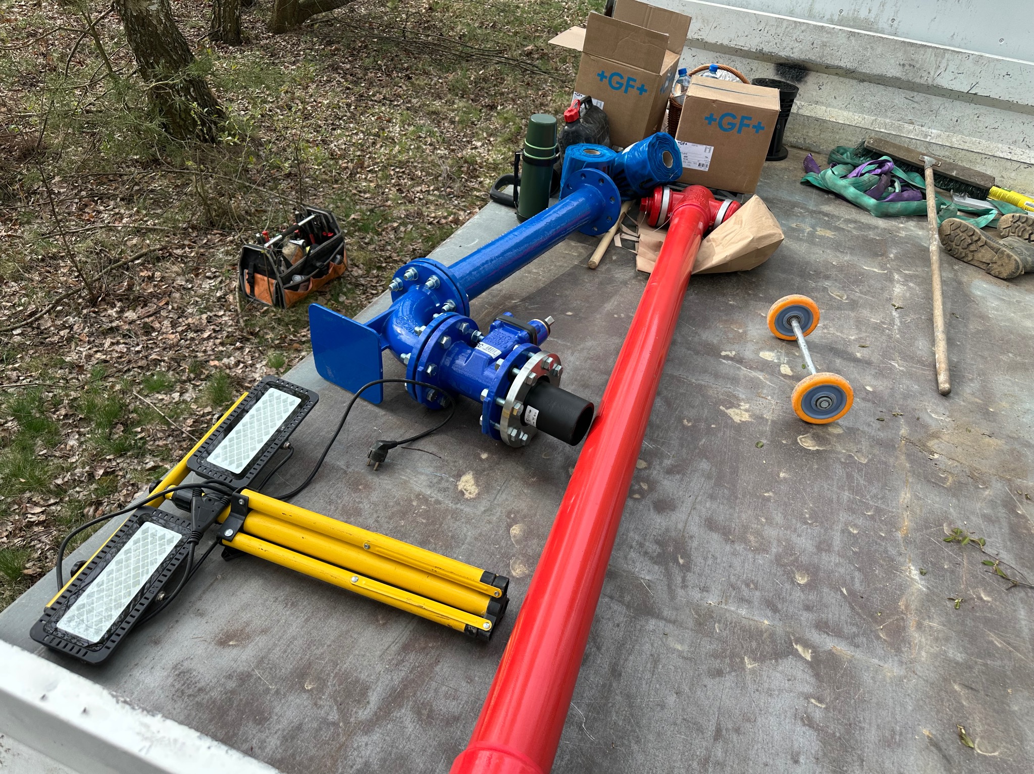 Elementy instalacji wodociągowej i gazowej przygotowane do montażu na pace samochodu, widoczne rury w kolorze czerwonym i niebieskim, skrzynka narzędziowa, lampa budowlana, oraz kartony z logo...