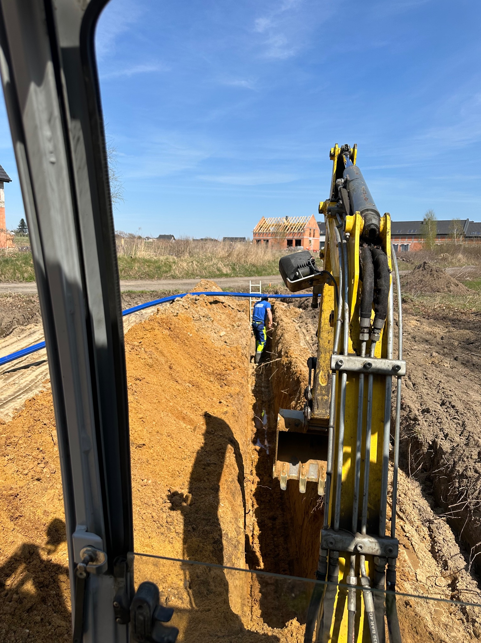 Widok z kabiny koparki na wykop z ułożonymi niebieskimi rurami, pracownik w oddali w odblaskowym ubraniu, w tle budowa domu w stanie surowym.