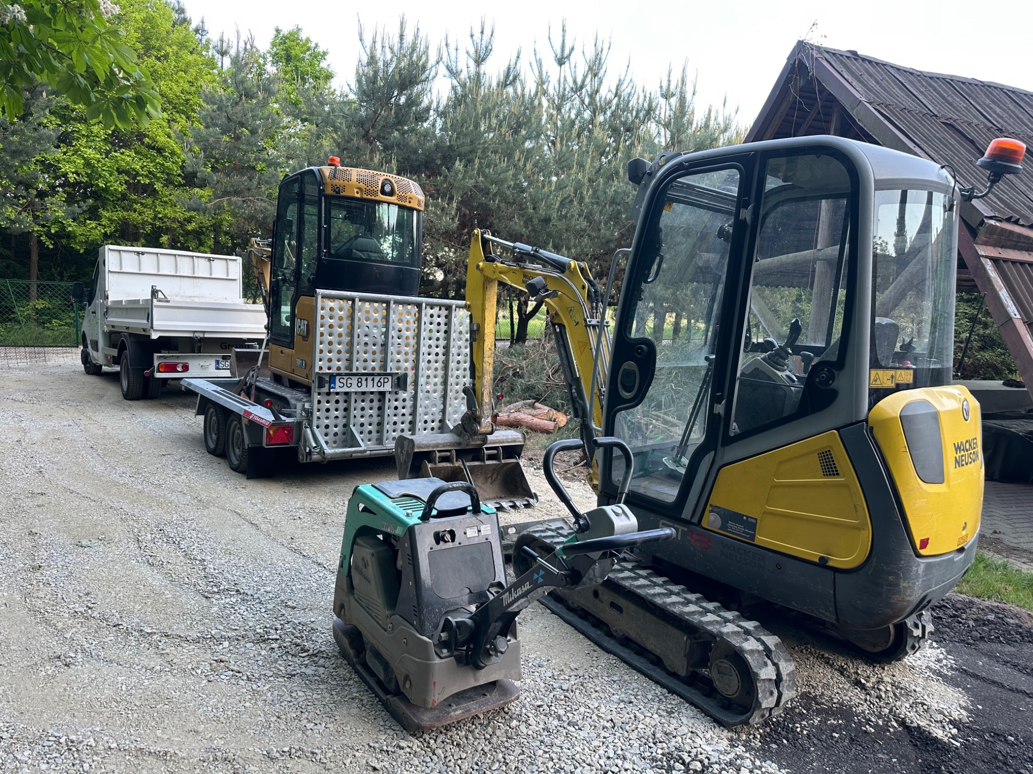 Żółta mini koparka Wacker Neuson i zagęszczarka Mikasa stoją na żwirowym podłożu. W tle widoczny transport z drugą koparką i drzewa.
