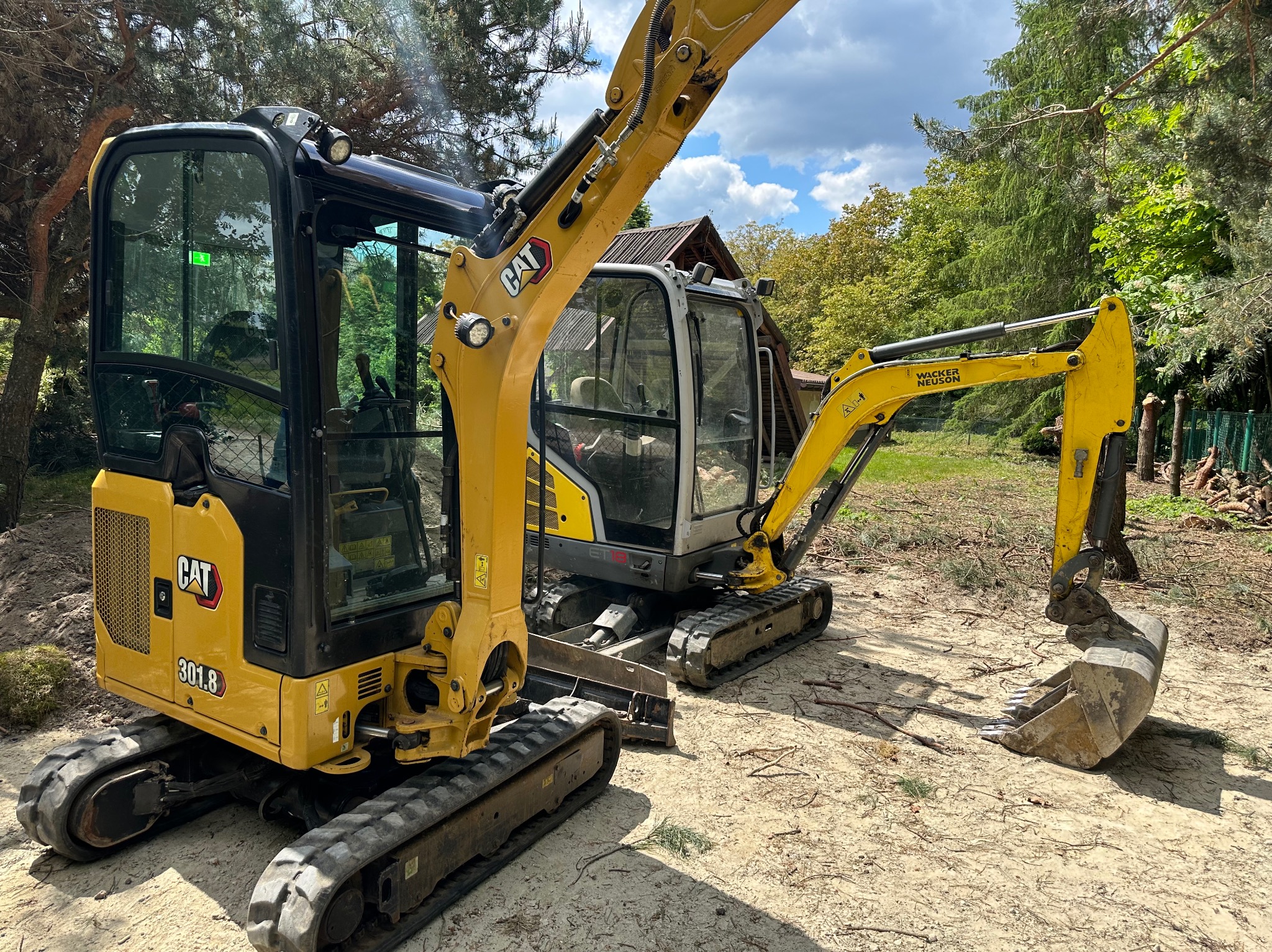 Dwie mini koparki: żółta CAT 301.8 z widocznym logo i szara Wacker Neuson, stoją na piaszczystym terenie z zielenią w tle, gotowe do pracy.