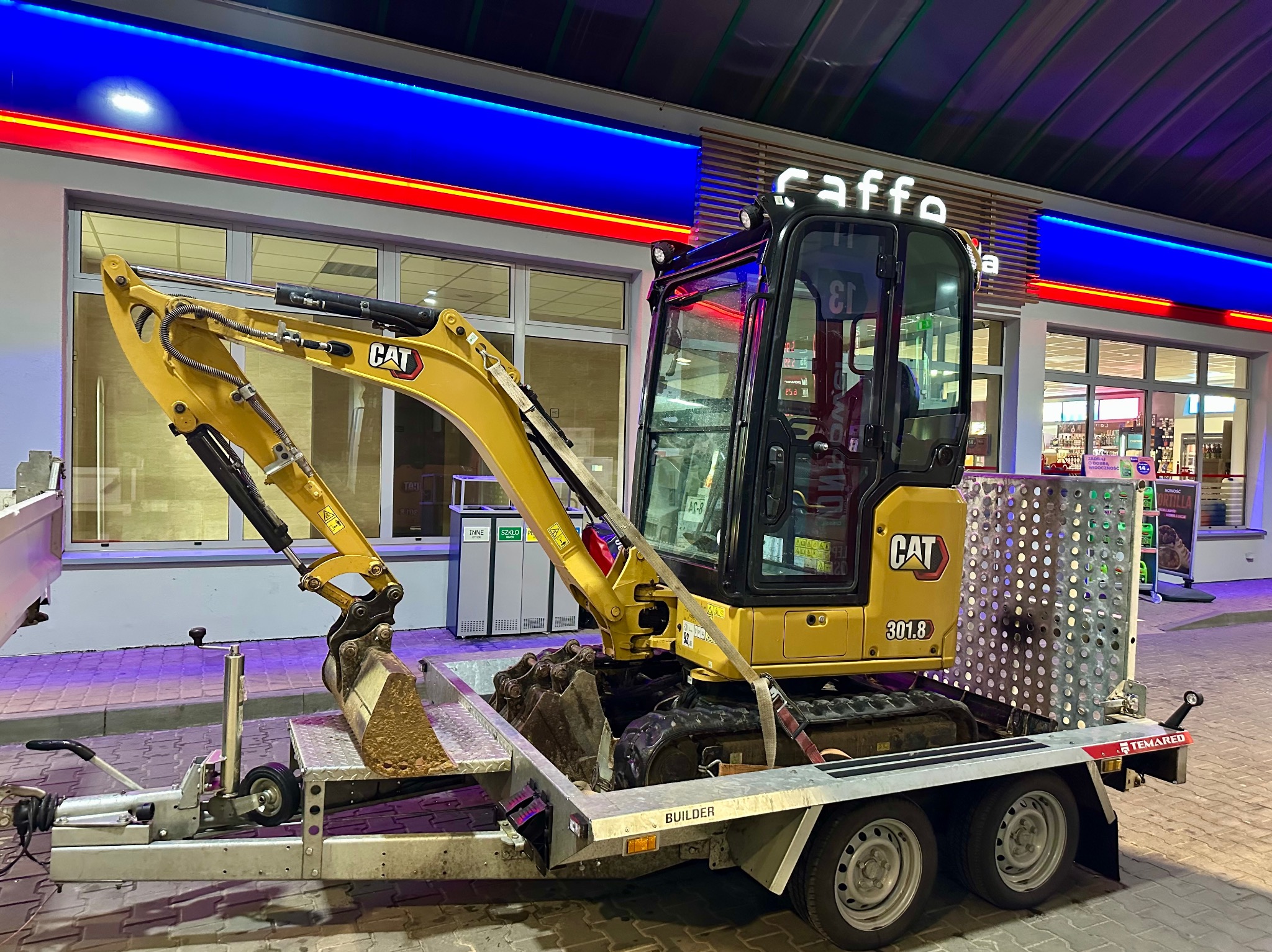 Żółta mini koparka CAT 301.8 na przyczepie, z łyżką lekko zabrudzoną ziemią. Tło stanowi budynek stacji benzynowej Caffe. Zdjęcie nocne, oświetlenie sztuczne.