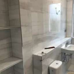 Nowoczesna łazienka w trakcie wykańczania, widoczne białe płytki imitujące marmur, wisząca umywalka, toaleta, półki w zabudowie, narzędzia i kable na blacie.