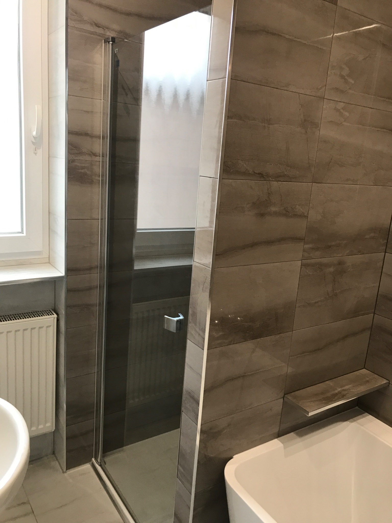 Nowoczesna kabina prysznicowa ze szklanymi drzwiami w łazience wykończonej płytkami imitującymi kamień, widoczna umywalka i fragment wanny.