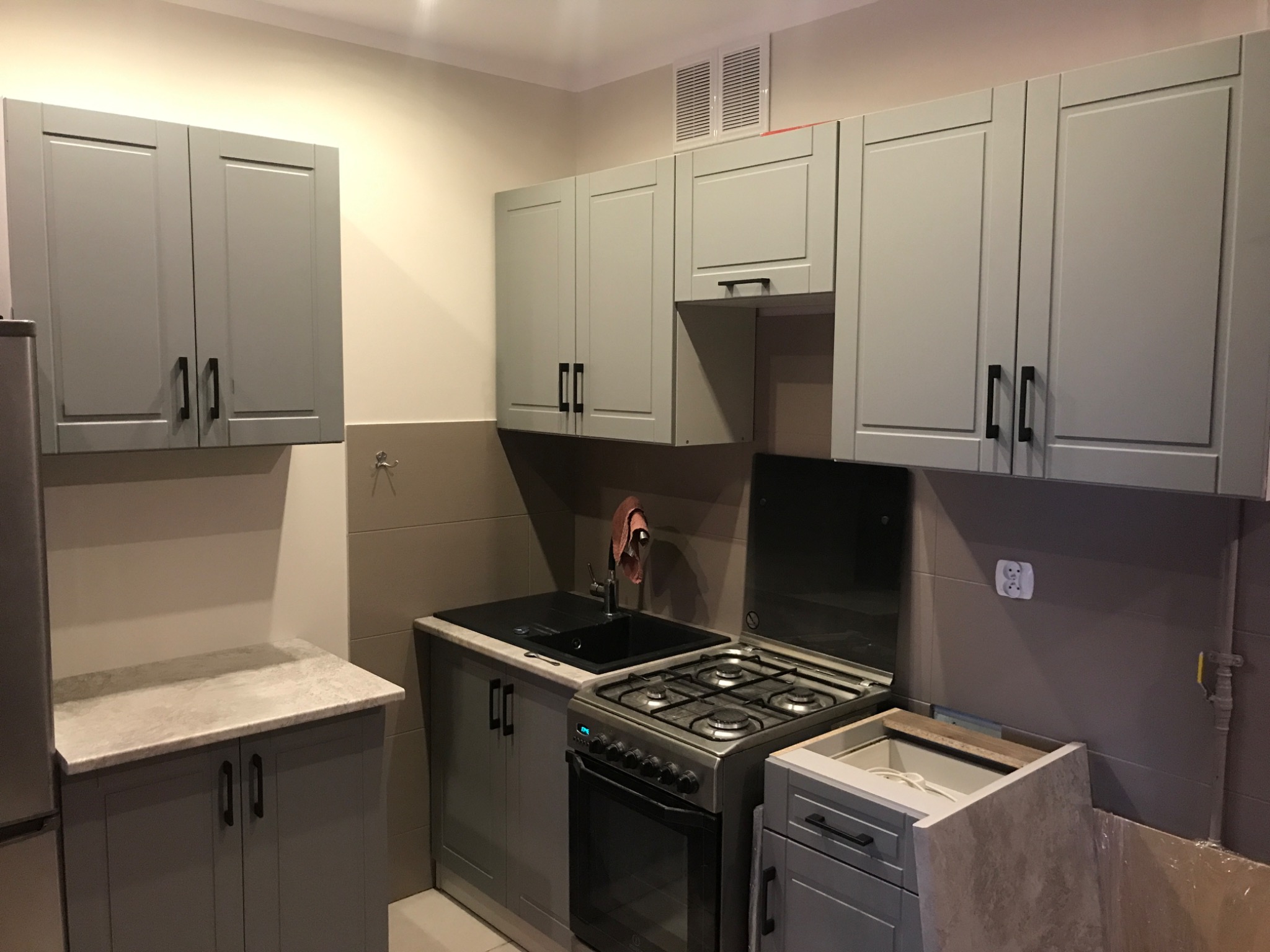 Szara kuchnia z zabudową meblową, czarnym zlewem i płytą gazową, widoczna wentylacja sufitowa i niezabezpieczone kable, fragment blatu roboczego z szarym wzorem marmuru.